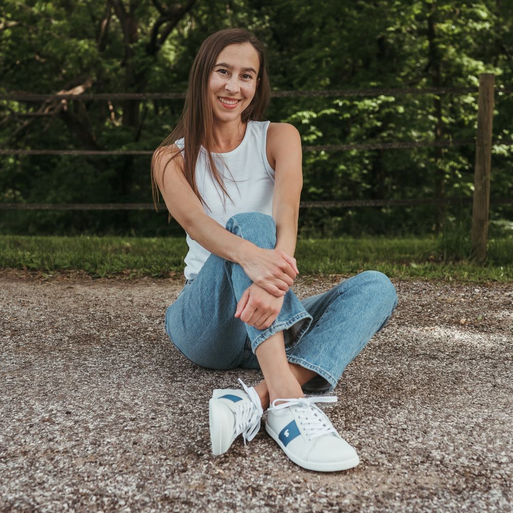 White leather Be Lenka Elite shoes with a thick, perforated, diagonal, blue leather stripe on both sides of the sneaker. A cursive, metal "be" logo design is in the center of the outside stripe. Both shoes are shown on a tan woman with long, brown hair wearing a white tank top tucked into light-wash loose-fitting blue jeans. She is sitting on a paved road with her legs loosely crossed and hands wrapped around her left knee leaning back a bit. #color_white-dark-blue