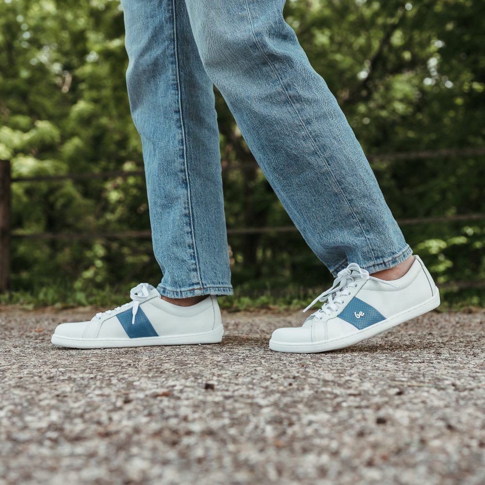 White leather Be Lenka Elite shoes with a thick, perforated, diagonal, blue leather stripe on both sides of the sneaker. A cursive, metal "be" logo design is in the center of the outside stripe. Both shoes are shown on a person wearing light-wash loose-fitting blue jeans facing left with their right foot stepping ahead of the left standing on a paved road with greenery in the background. #color_white-dark-blue