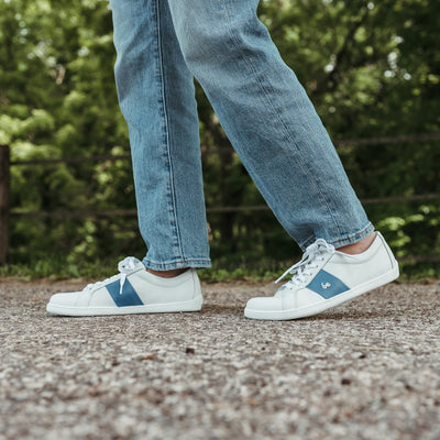 White leather Be Lenka Elite shoes with a thick, perforated, diagonal, blue leather stripe on both sides of the sneaker. A cursive, metal "be" logo design is in the center of the outside stripe. Both shoes are shown on a person wearing light-wash loose-fitting blue jeans facing left with their right foot stepping ahead of the left standing on a paved road with greenery in the background. #color_white-dark-blue