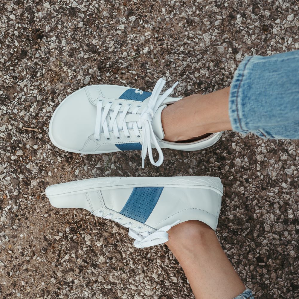 White leather Be Lenka Elite shoes with a thick, perforated, diagonal, blue leather stripe on both sides of the sneaker. A cursive, metal "be" logo design is in the center of the outside stripe. Both shoes are shown from above on a person wearing light-wash loose-fitting blue jeans sitting on a paved road. Left shoe and foot are tilted to the left, while the right foot is shown from above. #color_white-dark-blue