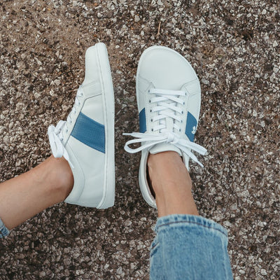 White leather Be Lenka Elite shoes with a thick, perforated, diagonal, blue leather stripe on both sides of the sneaker. A cursive, metal "be" logo design is in the center of the outside stripe. Both shoes are shown from above on a person wearing light-wash loose-fitting blue jeans sitting on a paved road. Left shoe and foot are tilted to the left, while the right foot is shown from above. #color_white-dark-blue