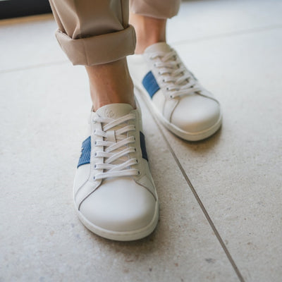 White leather Be Lenka Elite shoes with a thick, perforated, diagonal, blue leather stripe on both sides of the sneaker. A cursive, metal "be" logo design is in the center of the outside stripe. Both shoes are facing the camera on a person wearing rolled beige slacks standing on a large tile floor. #color_white-dark-blue