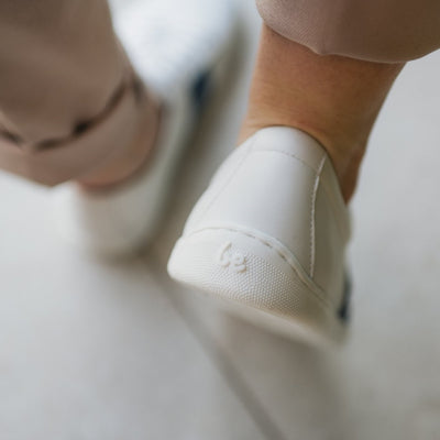 White leather Be Lenka Elite shoes with a thick, perforated, diagonal, blue leather stripe on both sides of the sneaker. A cursive, metal "be" logo design is in the center of the outside stripe. Right shoe heel is accented here on a person wearing rolled beige slacks standing on a large tile floor. #color_white-dark-blue