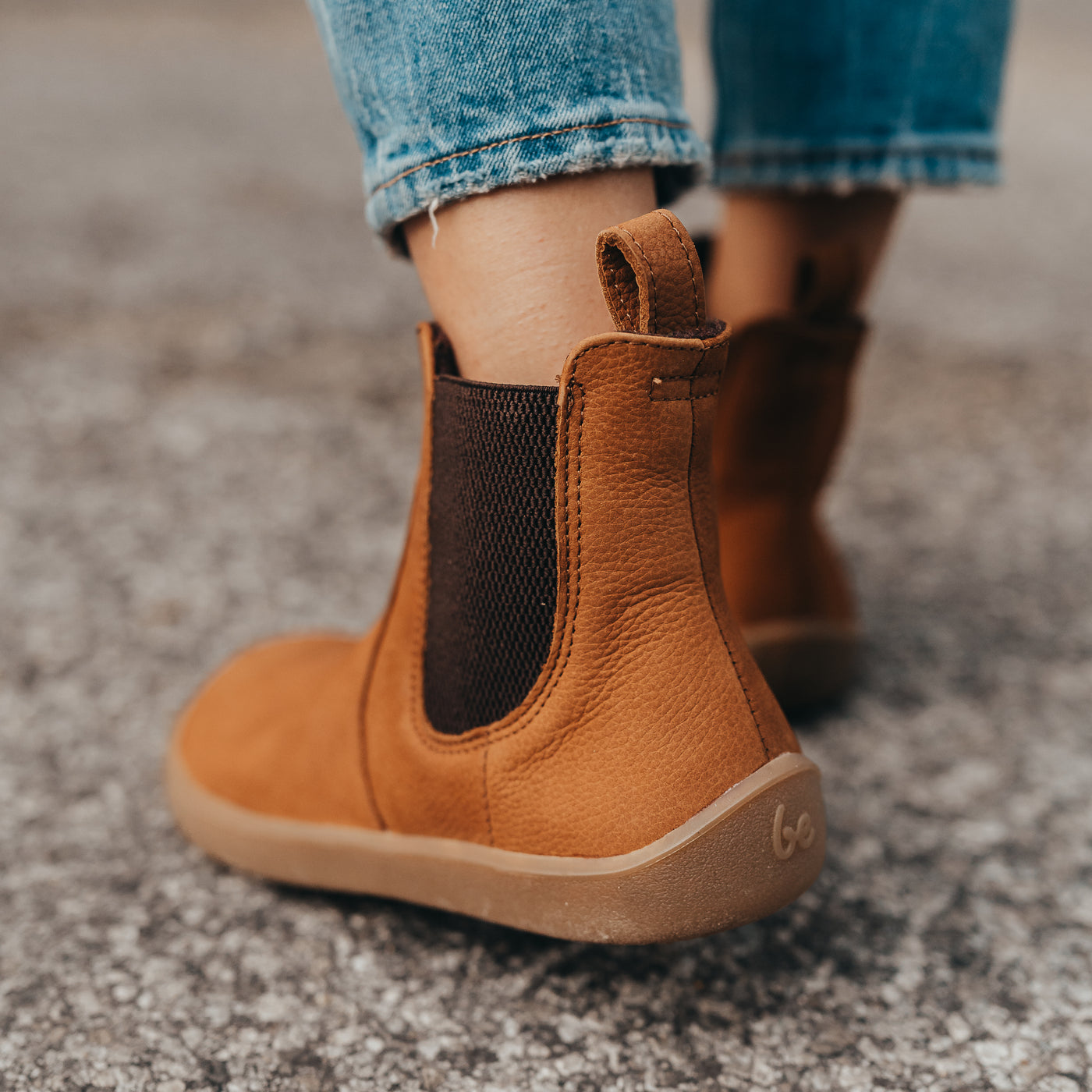 A photo of Be Lenka Entice boots made from nubuck leather and rubber soles. The boots are cinnamon in color, they are a Chelsea boot style with elastic at the sides. Both boots are shown on a woman's feet from behind with a close up of the back left side and heel of the left boot. The woman is wearing cropped blue skinny jeans and is walking on an asphalt road with her right foot in front of her left. #color_cinnamon