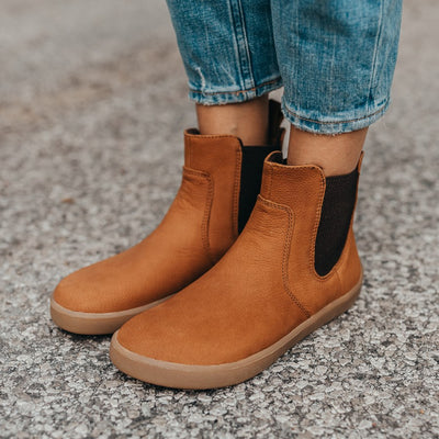 A photo of Be Lenka Entice boots made from nubuck leather and rubber soles. The boots are cinnamon in color, they are a Chelsea boot style with elastic at the sides. Both boots are shown on a woman's feet from the front left side. The woman is wearing cropped blue jeans and is standing with her feet together on an asphalt road. #color_cinnamon