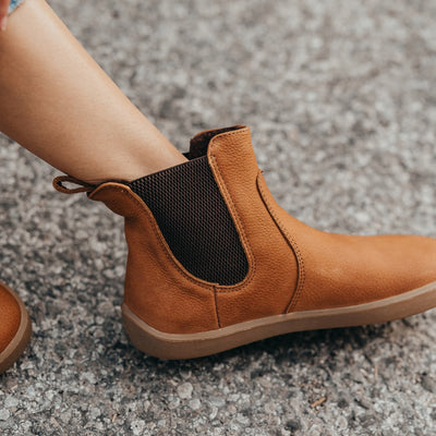 A photo of Be Lenka Entice boots made from nubuck leather and rubber soles. The boots are cinnamon in color, they are a Chelsea boot style with elastic at the sides. The boots are worn by a woman who is displaying the right side of the left boot and her bare shin on an asphalt road. #color_cinnamon
