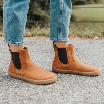 A photo of Be Lenka Entice boots made from nubuck leather and rubber soles. The boots are cinnamon in color, they are a Chelsea boot style with elastic at the sides. Both boots are shown on a woman's feet from the right side. The woman is wearing cropped blue jeans and is standing with her left foot in front of her right on an asphalt road with green grass in the background. #color_cinnamon