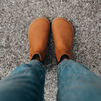 A photo of Be Lenka Entice boots made from nubuck leather and rubber soles. The boots are cinnamon in color, they are a Chelsea boot style with elastic at the sides. Both boots are shown on a woman's feet from above. The woman is wearing cropped blue skinny jeans and is standing on an asphalt road with her feet together. #color_cinnamon