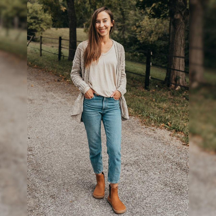 A photo of Be Lenka Entice boots made from nubuck leather and rubber soles. The boots are cinnamon in color, they are a Chelsea boot style with elastic at the sides. Both boots are shown on a woman's feet from the front. The woman has brown hair and is wearing cropped blue skinny jeans and a tan cable knit sweater over a tan shirt, and she is smiling while standing on an asphalt road with green grass, trees, and a black fence in the background. #color_cinnamonon
