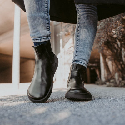A photo of Belenka Entice Neo boots made from smooth leather and black rubber soles. The boots are black in color with elastic panels on the sides and pull on loops. A woman is shown from the mid leg down standing on pavement and wearing gray skinny jeans and the boots. #color_black
