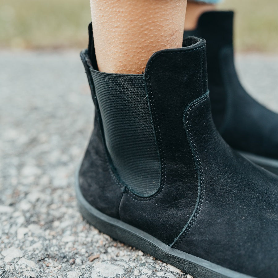 A photo of Be Lenka Entice boots made from nubuck leather and rubber soles. The boots are black in color, they are a Chelsea boot style with elastic at the sides. Both boots are shown on a woman's feet from the diagonal front right side with a close up of the elastic panel on the outer right boot. The woman is standing with her feet together on an asphalt road. #color_matte-black