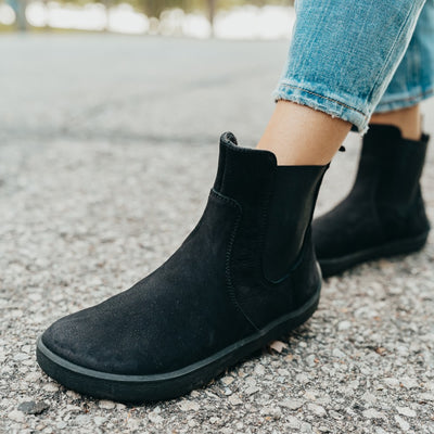 A photo of Be Lenka Entice boots made from nubuck leather and rubber soles. The boots are black in color, they are a Chelsea boot style with elastic at the sides. Both boots are shown on a woman's feet from the diagonal front left side. The woman is wearing blue cropped jeans and is standing with her left foot in front of her right on an asphalt road. #color_matte-black
