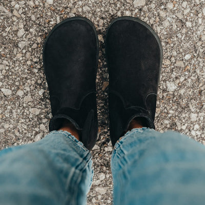 A photo of Be Lenka Entice boots made from nubuck leather and rubber soles. The boots are black in color, they are a Chelsea boot style with elastic at the sides. Both boots are shown on a woman's feet from above. The woman is wearing blue cropped jeans and is standing with her feet together on an asphalt road. #color_matte-black