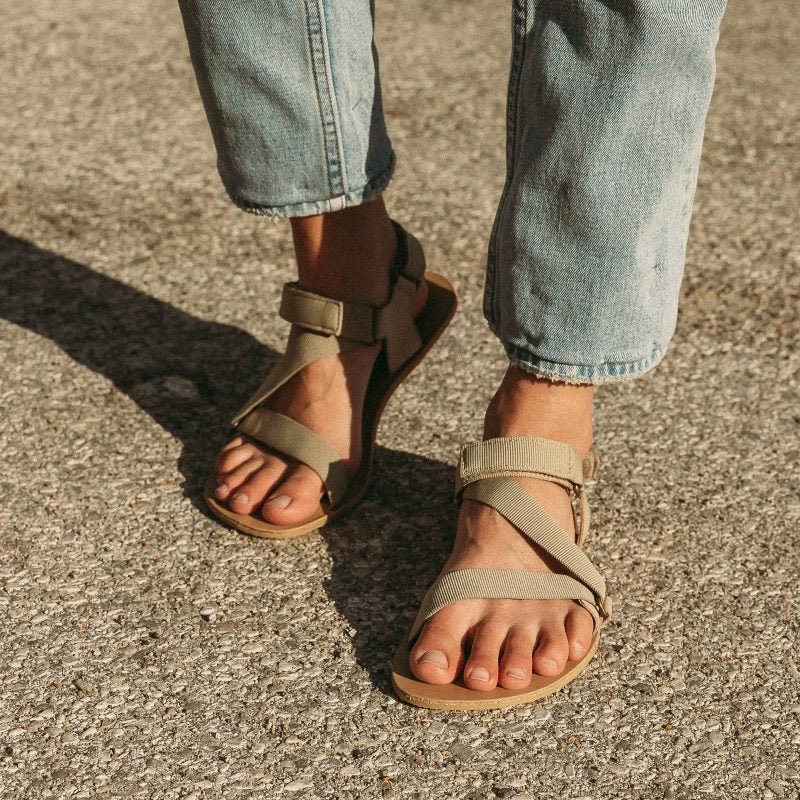 Be Lenka Flexi sandals in beige green. Sandals feature an adjustable fabric z-strap design. Soles are tan leather with tan rubber soles. Shoes are shown from the front on a womans feet wearing ankle length light blue jeans standing on grey pavement. #color_beige-green