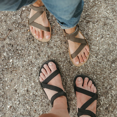 A photo of Black Be Lenka Flexi Sandals made with fabric straps with velcro and a tan leather topped rubber soles. The sandals are a black color and have straps that cross the front of the foot and continue around the mid-foot, ankle, and heel. Both sandals are shown from above on a light-skinned woman standing on pavement in front of another woman wearing the olive green colorway. #color_black