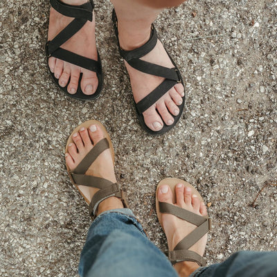 A photo of Olive Green Be Lenka Flexi Sandals made with fabric straps with velcro and a tan leather topped rubber soles. The sandals have straps that cross the front of the foot and continue around the mid-foot, ankle, and heel. Both shoes are shown crossed from above on a tan woman wearing rolled jeans standing on a paved road in front of a lighter-skinned woman wearing the black colorway. #color_olive-green