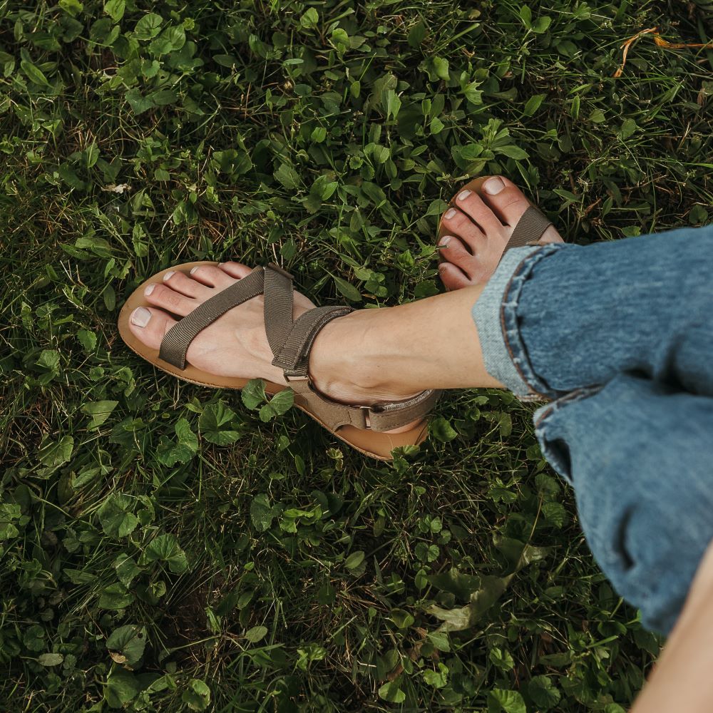 A photo of Olive Green Be Lenka Flexi Sandals made with fabric straps with velcro and a tan leather topped rubber soles. The sandals have straps that cross the front of the foot and continue around the mid-foot, ankle, and heel. Both shoes are shown crossed from above on a tan woman wearing rolled jeans sitting in grass. #color_olive-green