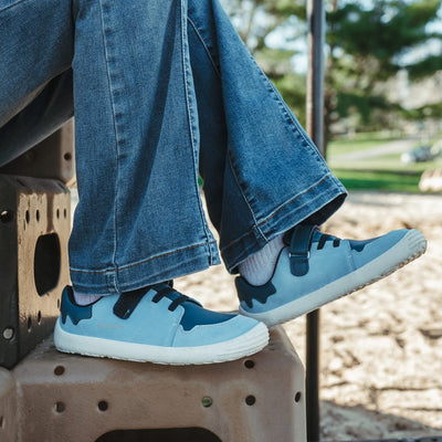 A photo of blue Be Lenka Gelato kids leather sneakers with white rubber soles. Shoes are light blue with dark blue accent color blocks on the tongue, toe box, single top velcro closure, two elastic laces below that, and a "dripping" ice cream color block on the heel. Shoes are shown facing right on a young girl wearing flare jeans sitting on playground stairs with a playground in the background. #color_blue