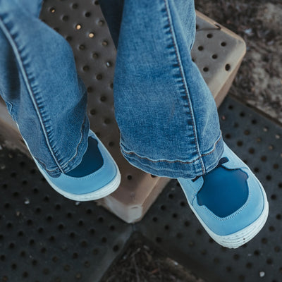 A photo of blue Be Lenka Gelato kids leather sneakers with white rubber soles. Shoes are light blue with dark blue accent color blocks on the tongue, toe box, single top velcro closure, two elastic laces below that, and a "dripping" ice cream color block on the heel. Shoes are shown from above on a young girl wearing flare jeans sitting on playground stairs. #color_blue