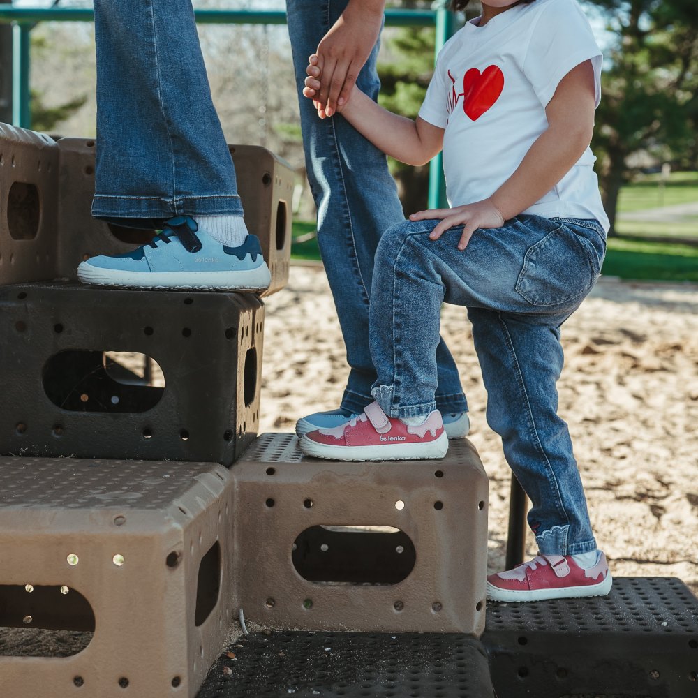 A photo of blue Be Lenka Gelato kids leather sneakers with white rubber soles. Shoes are light blue with dark blue accent color blocks on the tongue, toe box, single top velcro closure, two elastic laces below that, and a "dripping" ice cream color block on the heel. Shoes are shown facing left on a young girl wearing flare jeans standing on playground stairs holding hands with a little girl wearing pink Gelato shoes. #color_blue