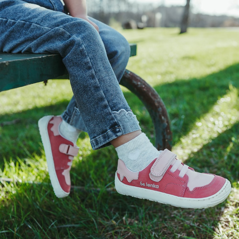 A photo of Pink Be Lenka Gelato kids leather sneakers with white rubber soles. Shoes are dark pink with light pink accent color blocks on the tongue, toe box, single top velcro closure, two elastic laces below that, and a "dripping" ice cream color block on the heel. Shoes are shown on a little girl wearing jeans sitting on a picnic bench kicking her right foot out with grass in the background. #color_pink