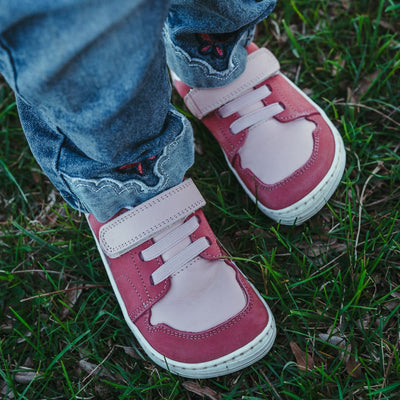 A photo of Pink Be Lenka Gelato kids leather sneakers with white rubber soles. Shoes are dark pink with light pink accent color blocks on the tongue, toe box, single top velcro closure, two elastic laces below that, and a "dripping" ice cream color block on the heel. Shoes are shown from above on a little girl wearing jeans standing in grass. #color_pink
