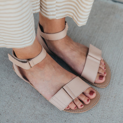 A photo of beige leather Be Lenka Iris Sandals. An adjustable strap wraps around the ankle once with an open heel while a thicker adjustable strap goes over the toes. A woman is shown from mid leg down wearing a striped skirt and the iris sandals, her feet are facing diagonally standing on concrete with a block wall in the background. #color_nude