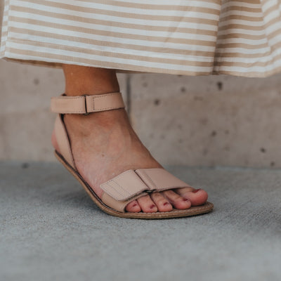 A photo of beige leather Be Lenka Iris Sandals. An adjustable strap wraps around the ankle once with an open heel while a thicker adjustable strap goes over the toes. A woman is shown from mid leg down wearing a striped skirt and the iris sandal, the heel of her foot is slightly lifted. She is standing on concrete with a block wall in the background. #color_nude