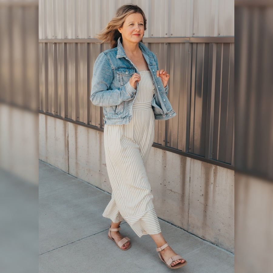 A photo of beige leather Be Lenka Iris Sandals. An adjustable strap wraps around the ankle once with an open heel while a thicker adjustable strap goes over the toes. Both sandals are shown on a woman’s feet. The woman is wearing a tan striped jumpsuit with a jean jacket and the Iris sandals, and she is walking on a paved sidewalk in front of an industrial building. #color_nude