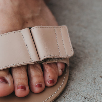A photo of beige leather Be Lenka Iris Sandals. An adjustable strap wraps around the ankle once with an open heel while a thicker adjustable strap goes over the toes. A woman is shown wearing the Iris sandal standing on concrete, the left sandal is shown up close from the front to show the detail of the velcro strap. #color_nude