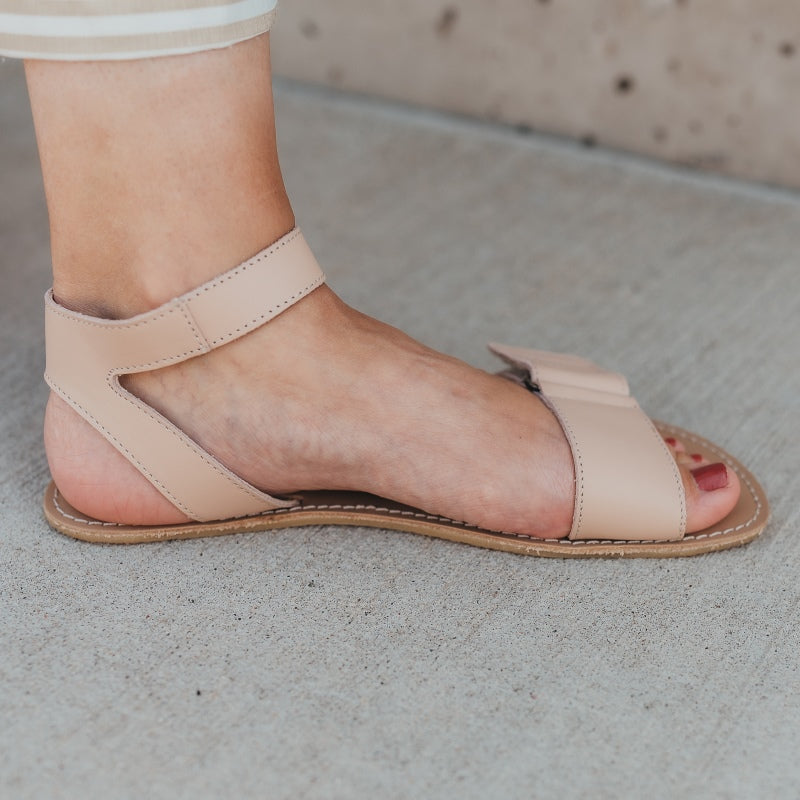 A photo of beige leather Be Lenka Iris Sandals. An adjustable strap wraps around the ankle once with an open heel while a thicker adjustable strap goes over the toes. A woman is shown from mid leg down wearing a striped skirt and the iris sandal, her foot is shown from the left side standing on concrete with a block wall in the background. #color_nude