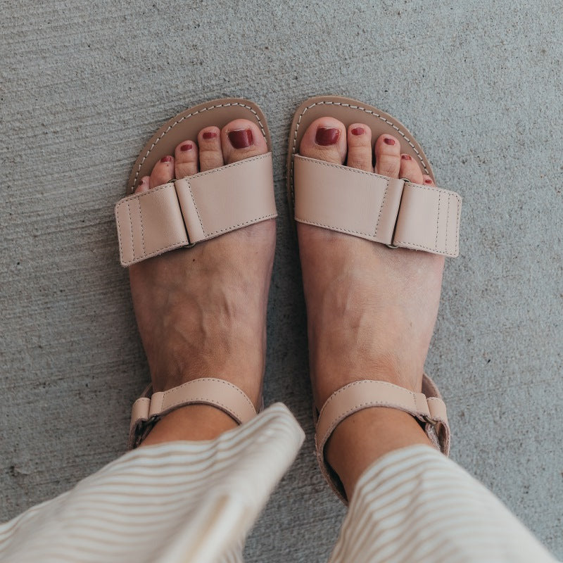 A photo of beige leather Be Lenka Iris Sandals. An adjustable strap wraps around the ankle once with an open heel while a thicker adjustable strap goes over the toes. A woman is shown from mid leg down wearing a striped skirt and the iris sandals, her feet are shown from the top down standing on concrete with a block wall in the background. #color_nude
