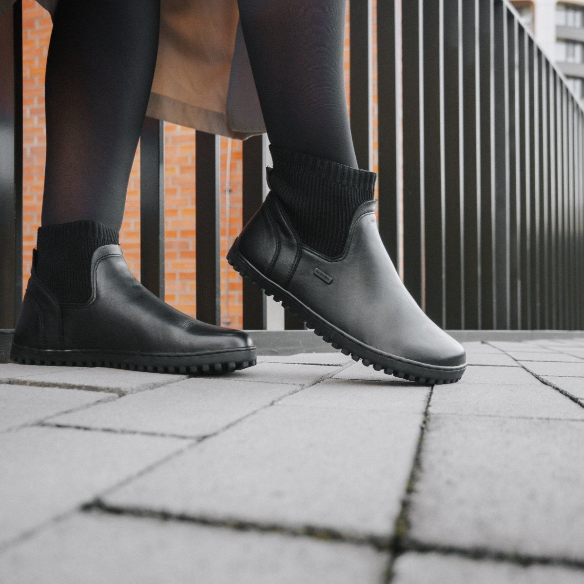 A photo of Be Lenka Mojo boots in black. Boots are made from smooth black leather and a knit opening hugging around the ankle. Both boots are shown from the right on a woman's feet. The woman is wearing black stockings and is walking on a paved sidewalk with a black metal fence in the background #color_black