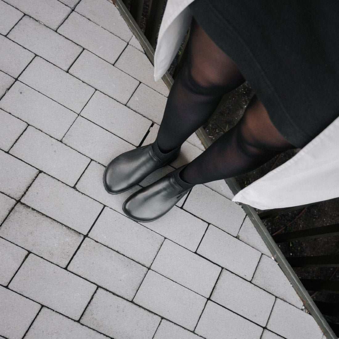 A photo of Be Lenka Mojo boots in black. Boots are made from smooth black leather and a knit opening hugging around the ankle. Both boots are shown from above on a woman's feet. The woman is wearing black stockings and and black skirt is standing on a paved sidewalk. #color_black