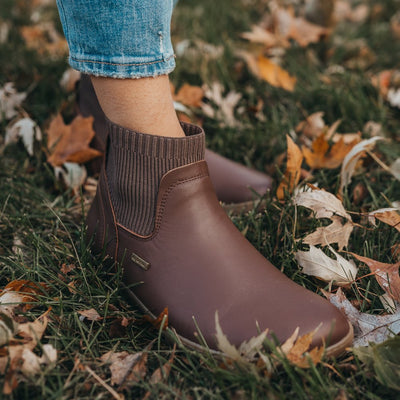 A photo of Be Lenka Mojo boots in dark brown with tan soles. Boots are made from smooth brown leather and a knit opening hugging around the ankle. Right boot is shown facing diagonally right on a woman wearing loose, distressed, cropped, light blue jeans standing in leafy grass with the left shoe behind the right. #color_dark-brown