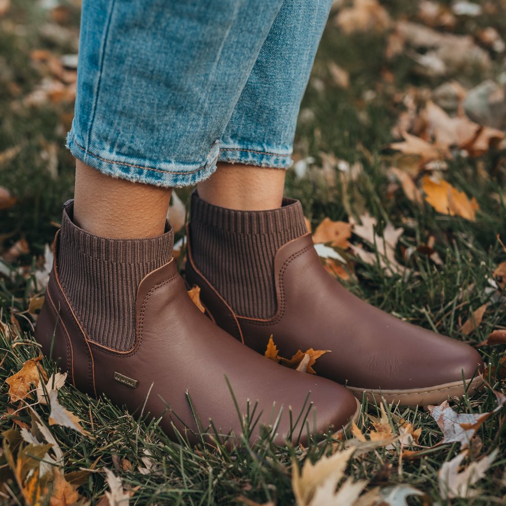 A photo of Be Lenka Mojo boots in dark brown with tan soles. Boots are made from smooth brown leather and a knit opening hugging around the ankle. Both shoes are shown facing right on a woman wearing loose, distressed, cropped, light blue jeans standing in leafy grass. #color_dark-brown