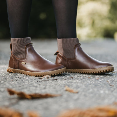 A photo of Be Lenka Mojo boots in dark brown with tan soles. Boots are made from smooth black leather and a knit opening hugging around the ankle. The shoes are shown from the right side on a woman's feet. The woman is wearing black stockings and is standing on a paved road with some fallen leaves in the foreground. #color_dark-brown