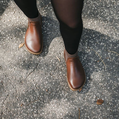 A photo of Be Lenka Mojo boots in dark brown with tan soles. Boots are made from smooth black leather and a knit opening hugging around the ankle. Both shoes are shown from above on a woman's feet. The woman is wearing black stockings and is walking on a paved road with some fallen leaves on it. #color_dark-brown