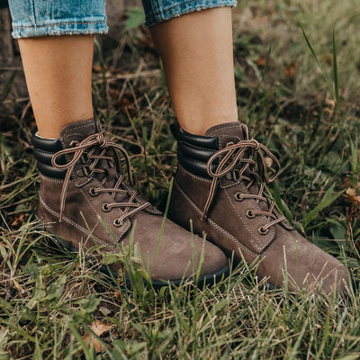 A photo of Be Lenka Nevada Boots made from nubuck leather and rubber soles. The boots are chocolate brown in color, they are combat boot style with laces a padded color and a chunky eyelets. Both boots are shown from the front right side on a woman's feet, with a view of her shins down. The woman is wearing cropped blue jeans and the boots and is standing in grass. #color_chocolate