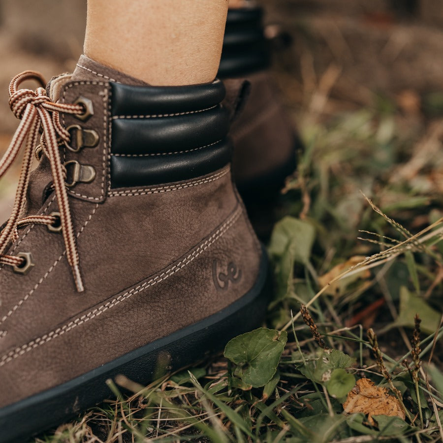 A photo of Be Lenka Nevada Boots made from nubuck leather and rubber soles. The boots are chocolate brown in color, they are combat boot style with laces a padded color and a chunky eyelets. The left boot is shown from the left side on a woman's feet, with a view of her ankles down with grass in the background. #color_chocolate