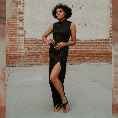 Black leather Be Lenka Promenade thong-style sandals with a thinner sling-back strap and a tan footbed and rubber soles. Both shoes are shown from the front on a tan woman with short, black, curly hair wearing a black, tank-top kimono-styled dress with a slit standing in a cement in front of a brick building. #color_black
