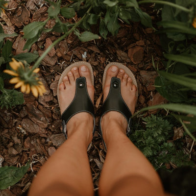 Black leather Be Lenka Promenade thong-style sandals with a thinner sling-back strap and a tan footbed and rubber soles. Both shoes are shown from above on a tan woman standing in a mulched garden. #color_black