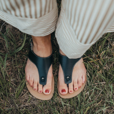 A photo of Black Be Lenka Promenade Sandals made with leather and tan rubber soles. The sandals have black thong straps and a heel strap with a buckle. A woman is shown standing in grass from mid leg down facing forward wearing the promenade sandals, a striped jumpsuit, and her toenails are painted red. #color_black