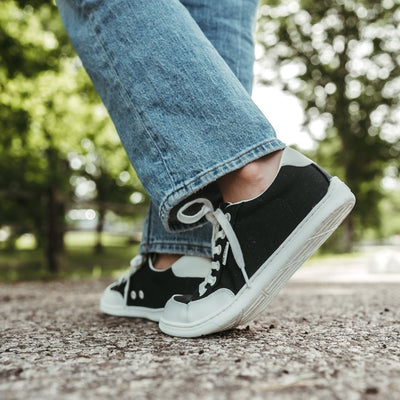 Black Be Lenka Rebound sneakers with white laces, microfiber toe guards, heel accents, and rubber soles. Both shoes are staggered shown facing back diagonally to the left on a woman wearing light-wash loose-fitting jeans on a paved road with greenery in the background. #color_black-white