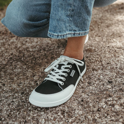 Black Be Lenka Rebound sneakers with white laces, microfiber toe guards, heel accents, and rubber soles. Left shoe is shown facing diagonally left on a woman wearing light-wash loose-fitting jeans kneeling on one knee on a paved road. #color_black-white