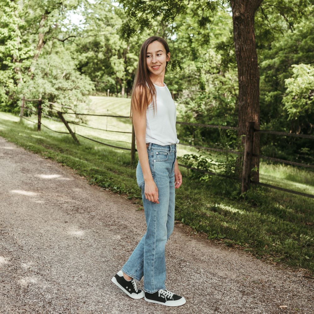 Black Be Lenka Rebound sneakers with white laces, microfiber toe guards, heel accents, and rubber soles. Both shoes are staggared facing right on a tan woman with long brown hair wearing light-wash loose-fitting jeans with a loose white tank top tucked in standing on a paved stone road with greenery in the background. #color_black-white