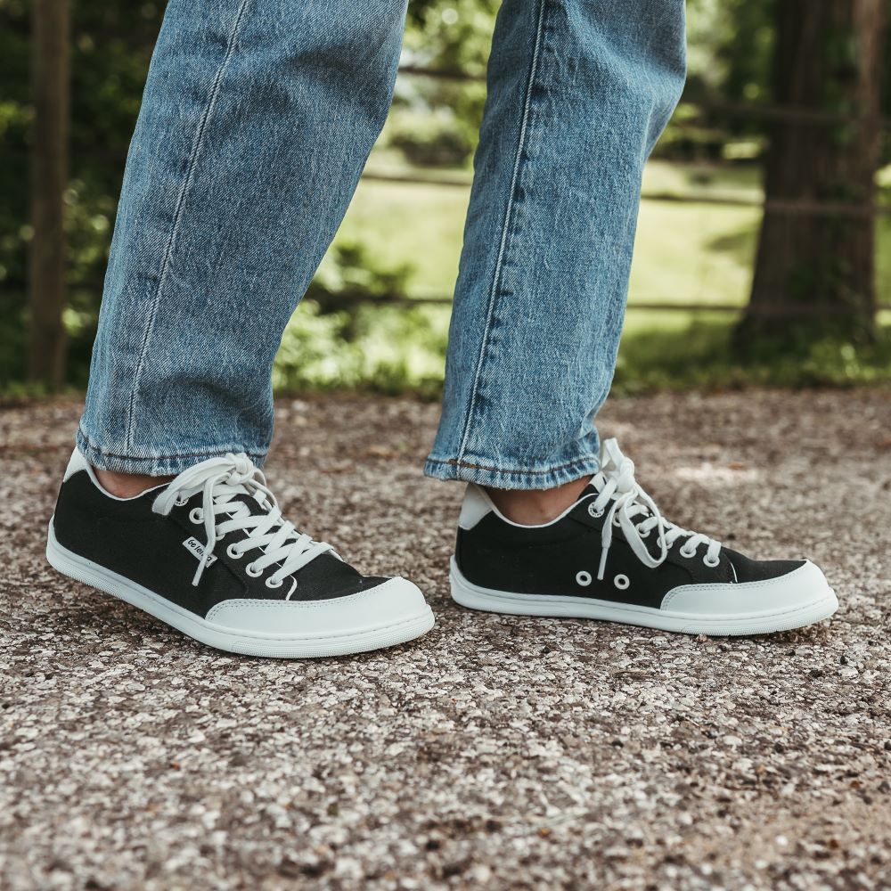 Black Be Lenka Rebound sneakers with white laces, microfiber toe guards, heel accents, and rubber soles. Both shoes are staggared facing right on a woman wearing light-wash loose-fitting jeans standing on a paved stone road with greenery in the background. #color_black-white