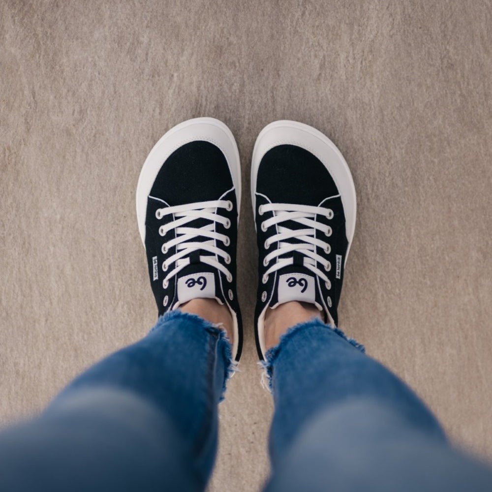 Black Be Lenka Rebound sneakers with white laces, microfiber toe guards, heel accents, and rubber soles. Both shoes are shown from above on a woman wearing skinny blue jeans standing on a grey floor. #color_black-white