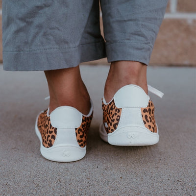 Leopard print Be Lenka Rebound sneakers with white laces, microfiber toe guards, heel accents, and rubber soles. Both shoes are shown from the back on a dark skinned woman wearing wide-leg, cropped, grey linen pants standing on cement with a brick wall in the background. #color_leopard