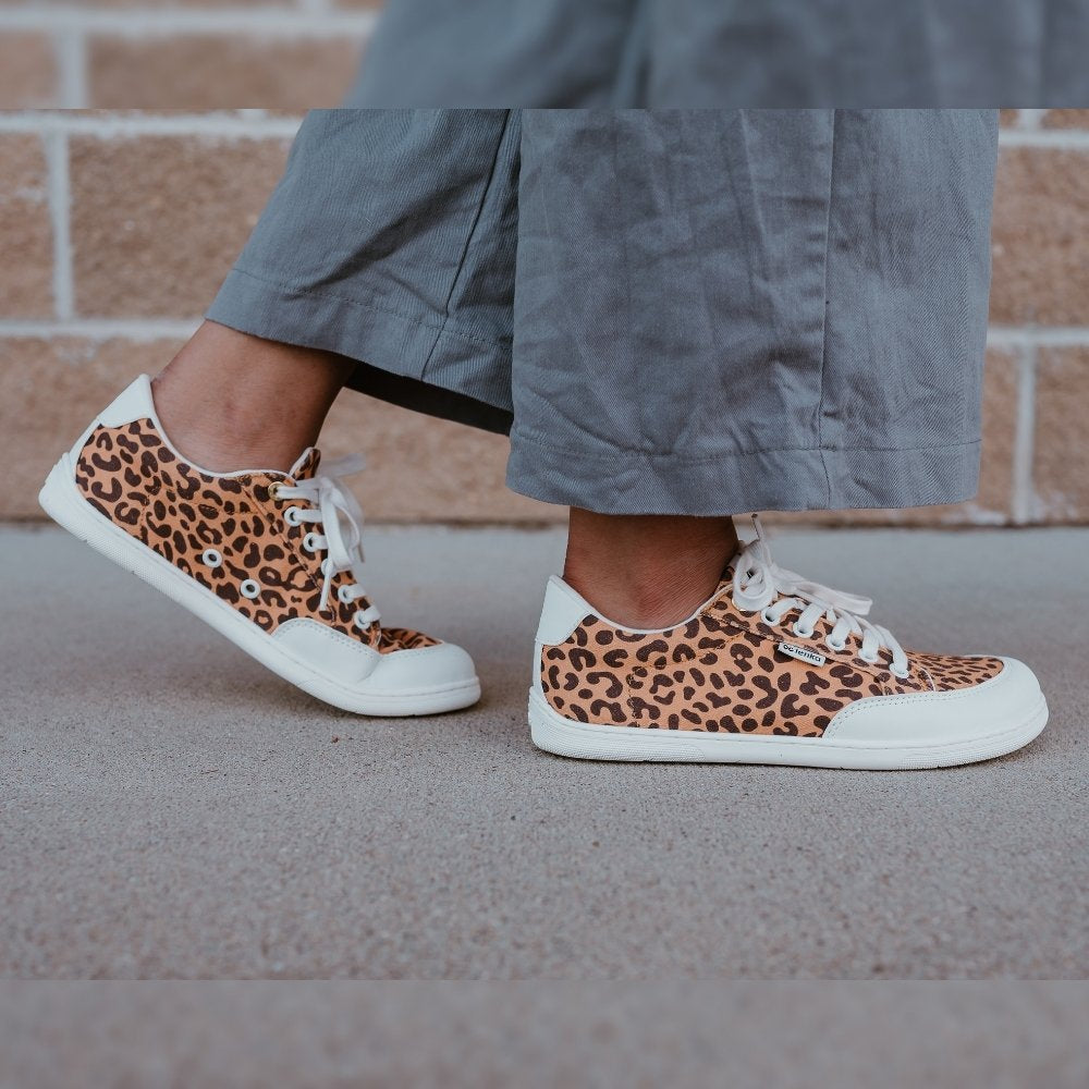 Leopard print Be Lenka Rebound sneakers with white laces, microfiber toe guards, heel accents, and rubber soles. Both shoes are shown staggered facing right on a dark skinned woman wearing wide-leg, cropped, grey linen pants standing on cement with a brick wall in the background. #color_leopard